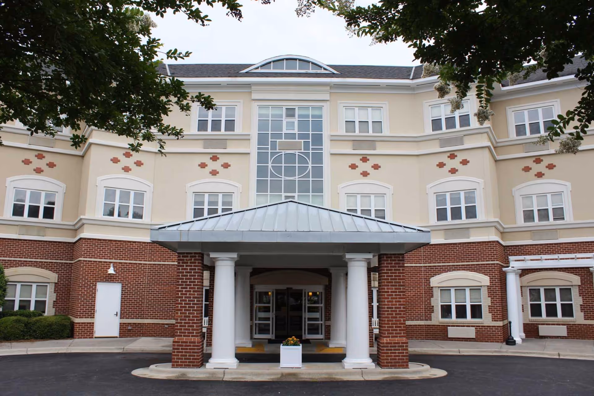 Front exterior view of a multi-story senior living facility building with a covered entrance supported by white columns, beige upper walls with decorative brick accents, and multiple windows. There are trees partially framing the top of the image.