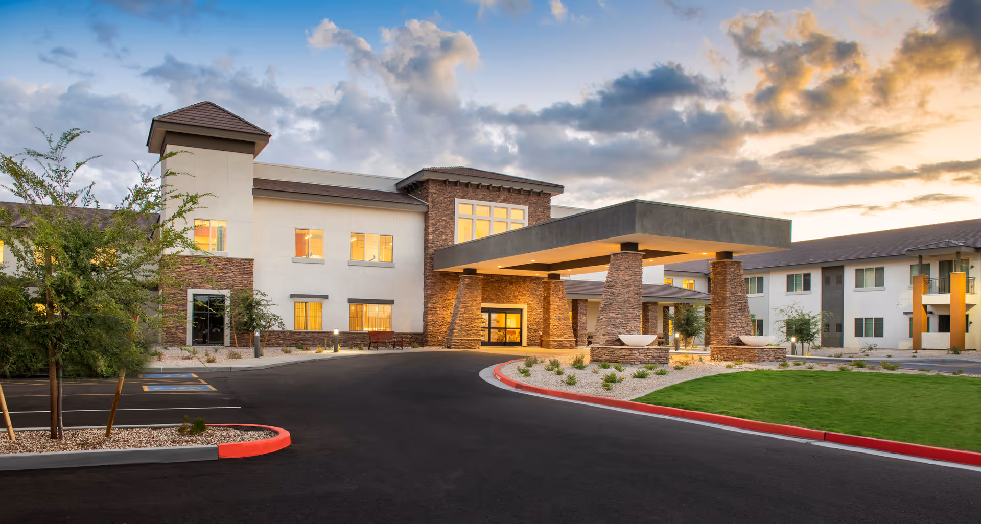 Exterior view of a modern senior living facility building at sunset with a covered entrance supported by stone pillars, landscaped greenery, and a paved driveway with red curbs.