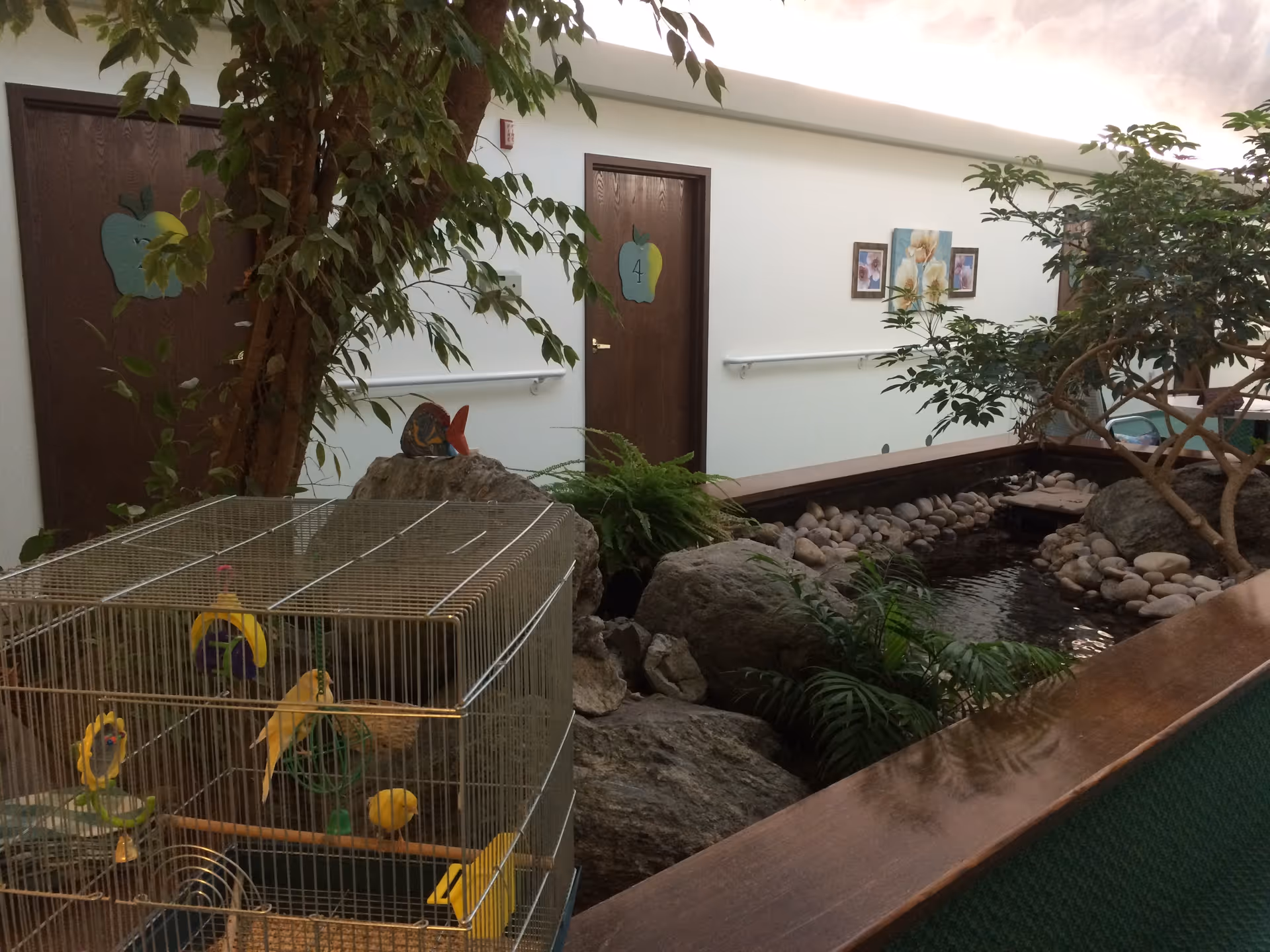 Indoor area of an assisted living facility featuring a small pond with rocks and plants, a birdcage with yellow birds, and several doors with apple-shaped signs numbered 4 and 5. The walls are decorated with framed pictures and there are handrails along the corridor.