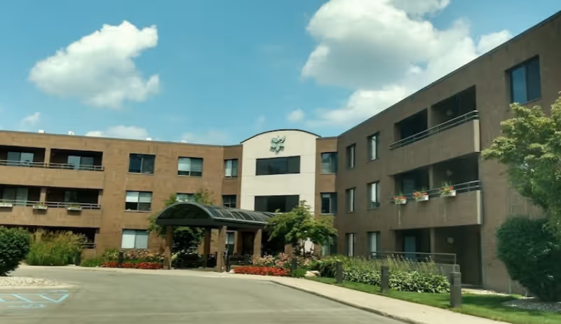 Exterior view of Robbinswood Assisted Living Community building with a covered entrance, three stories, balconies with flower pots, and landscaping including bushes and flowers under a partly cloudy sky.