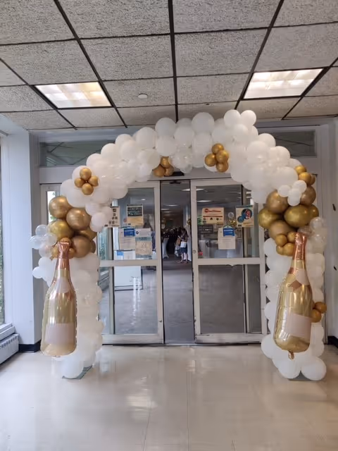 A decorative balloon arch made of white, gold, and transparent balloons with two large balloon champagne bottles on each side, positioned in front of glass double doors inside a building.