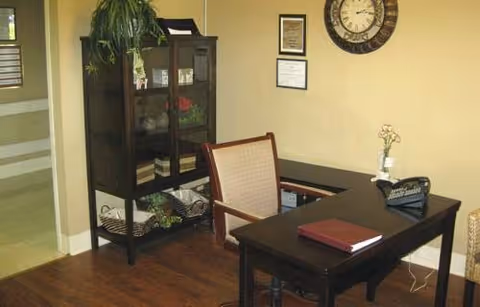 An interior office or reception area with a dark wooden desk and chair. On the desk are a closed book, a small flower vase, and a nameplate. Behind the desk is a glass-front cabinet with plants and decorative items. The wall has a large clock and framed certificates or documents. The floor is wooden, and there is a hallway visible to the left.