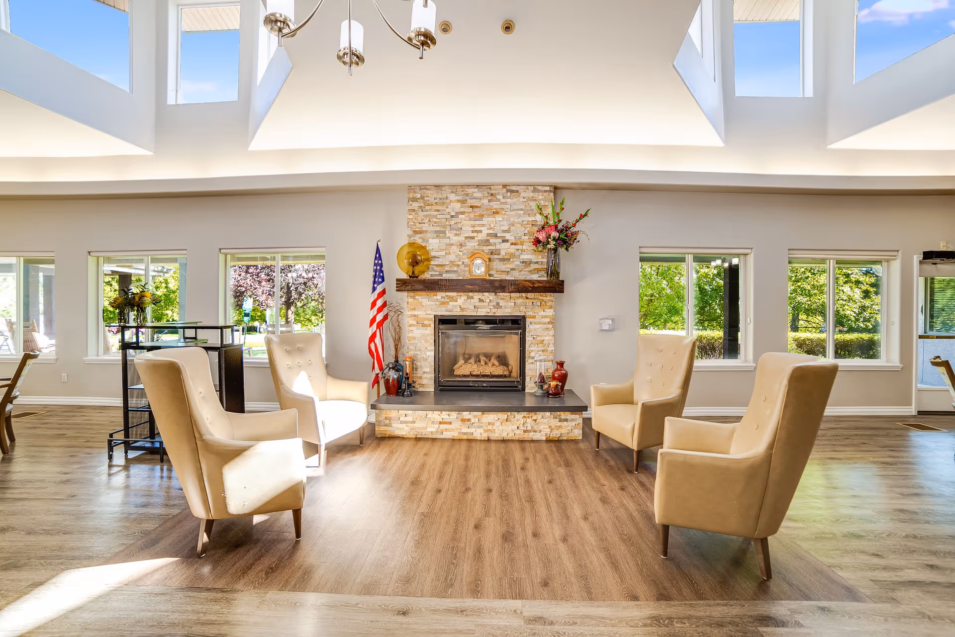 Sunlit communal lounge with a central stone fireplace, four beige armchairs, high skylit ceiling and large windows.