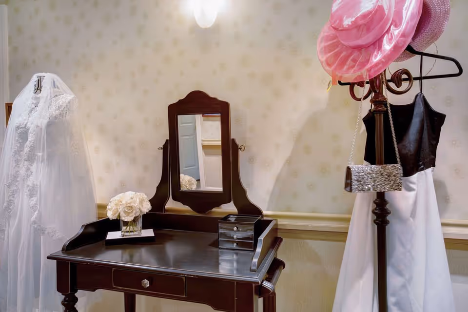 A vintage-style wooden vanity table with a mirror, a small jewelry box, and a vase of white flowers. To the left, a white lace wedding dress with a veil is displayed on a dress form. To the right, a black and white dress hangs on a coat rack along with two pink hats and a small silver handbag. The background features light-colored wallpaper with a subtle floral pattern.