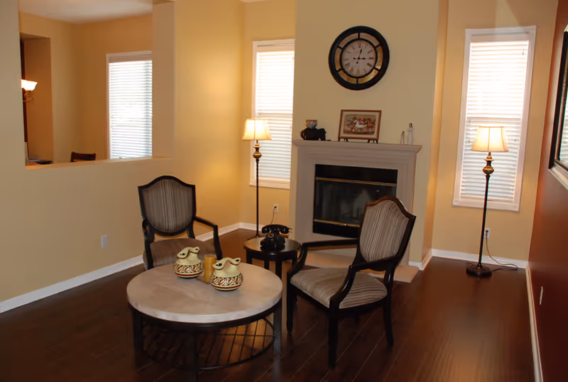 A cozy living room with two upholstered armchairs around a round coffee table with decorative pottery. The room features a fireplace with a clock and framed picture above it, two tall windows with blinds, and two floor lamps providing warm lighting. The walls are painted in beige and one side in a darker red tone, with dark wooden flooring.