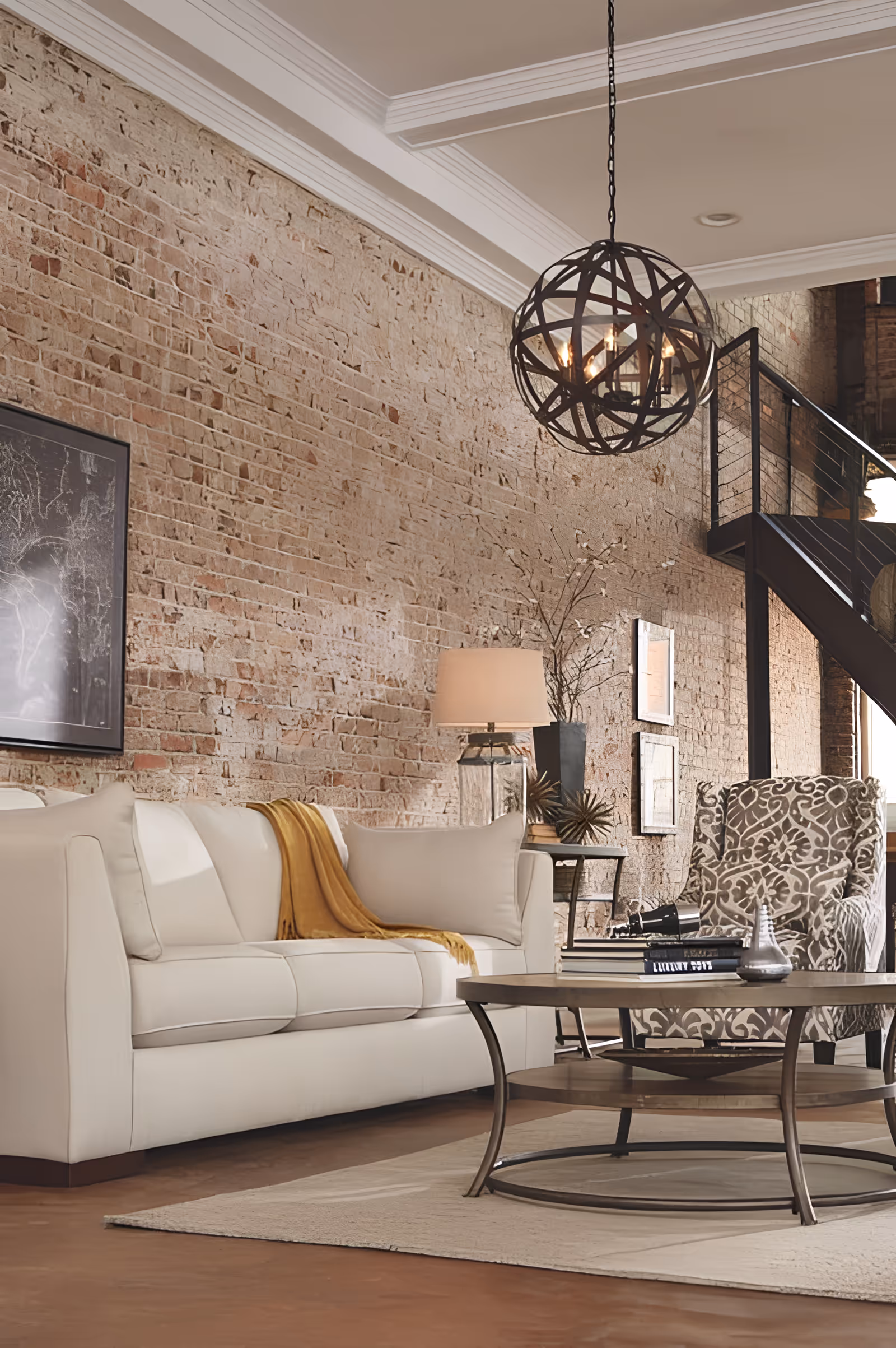A cozy living room with a white sofa draped with a mustard yellow throw blanket, a patterned armchair, and a round wooden coffee table with books and decorative items. The room features an exposed brick wall, a modern spherical chandelier, a table lamp, and a staircase with a metal railing in the background.