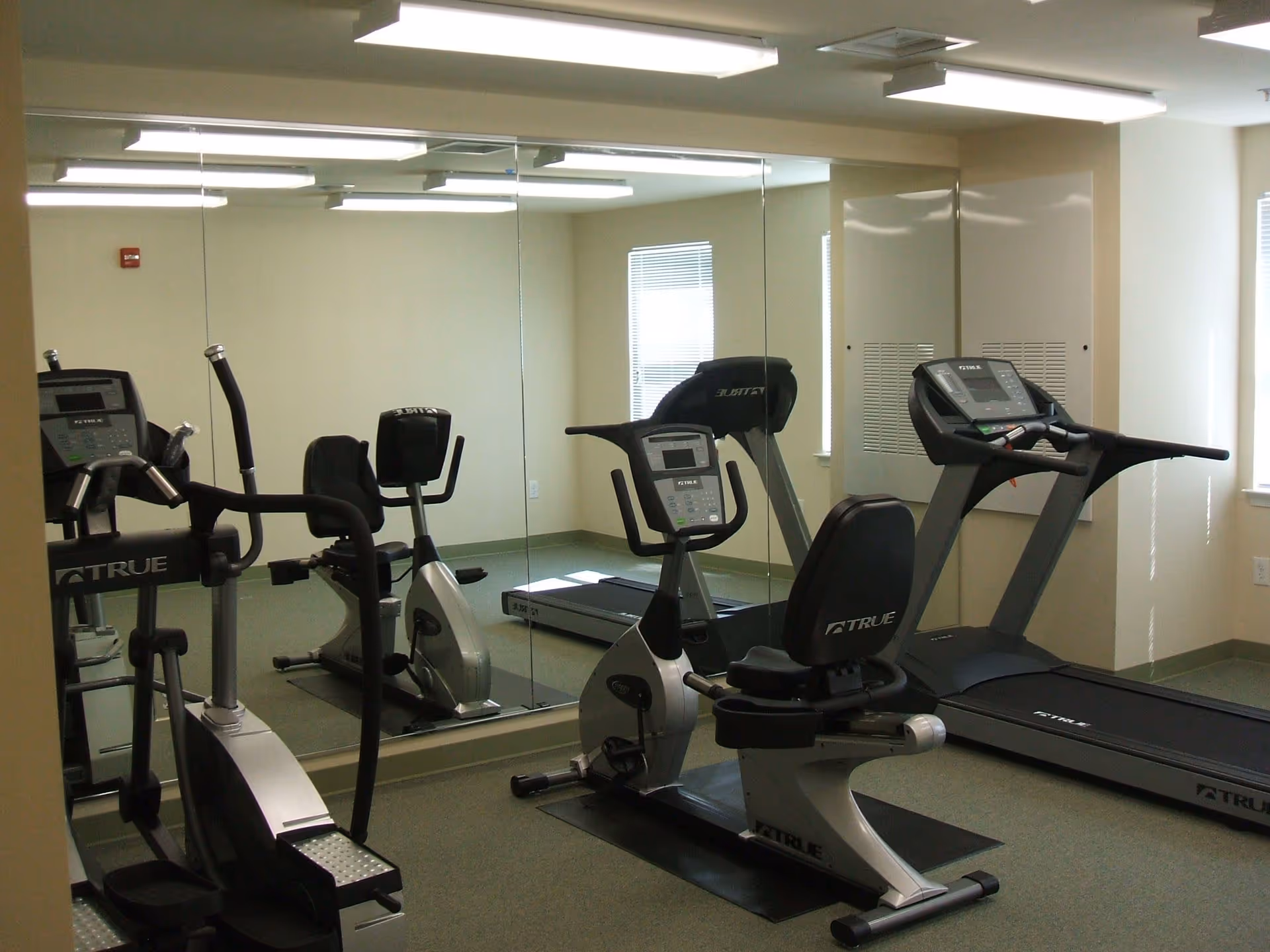 Small fitness room with exercise equipment including a recumbent stationary bike, treadmill, and elliptical machine. The room has beige walls, a large mirror on one wall, and windows letting in natural light.