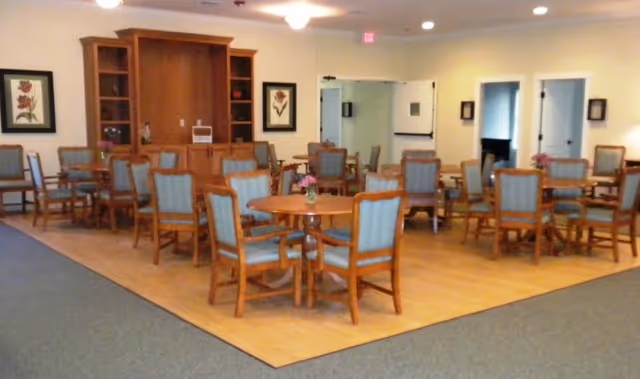 Dining/activity room with multiple round wooden tables and upholstered chairs on a wood floor and a cabinet against the wall.
