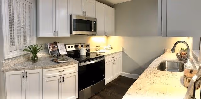 A modern kitchen with white cabinets, granite countertops, a stainless steel electric stove with oven, a microwave above the stove, a sink with a faucet on the right countertop, and decorative items including a small plant and a cookbook on the left countertop.
