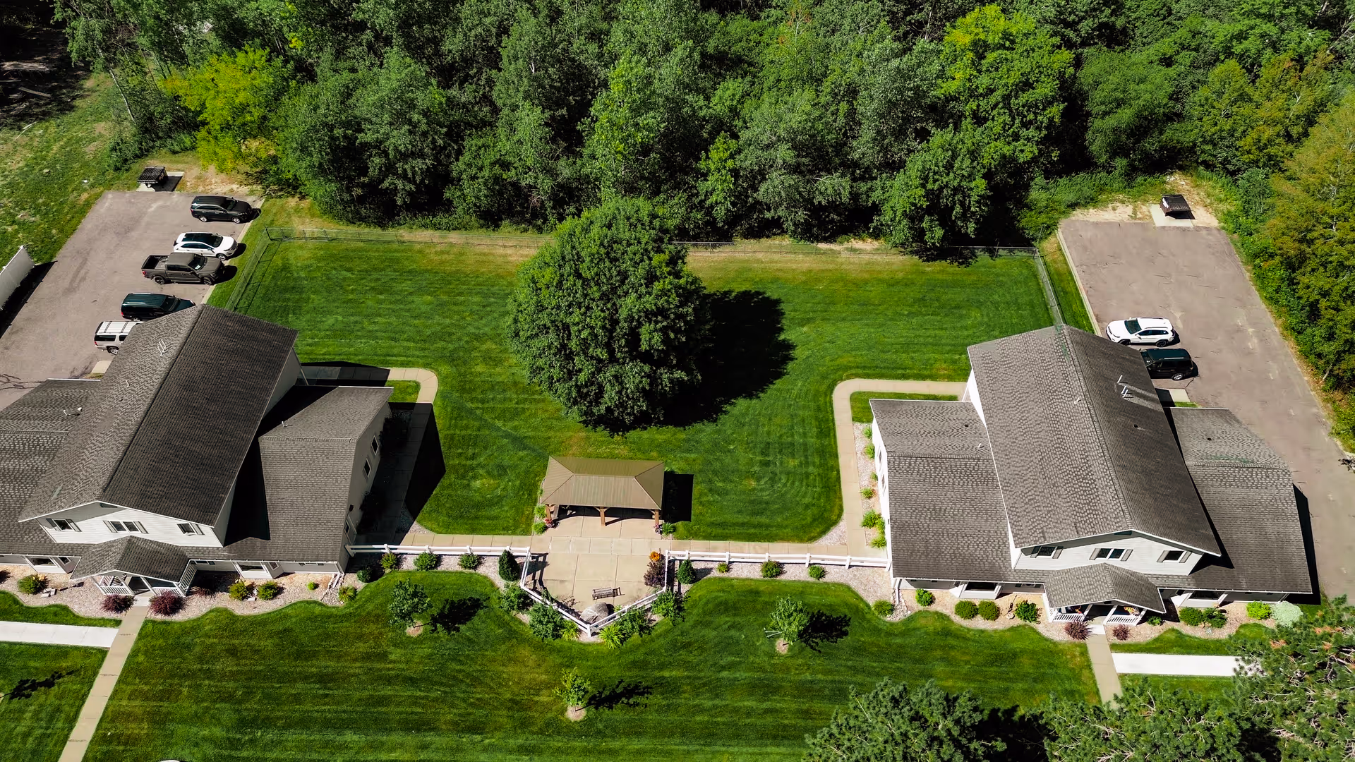Aerial view of Silver Maple Residence showing two large buildings separated by a green lawn with a central tree and a small gazebo. There are parking areas with several cars on either side of the buildings, and dense trees in the background.