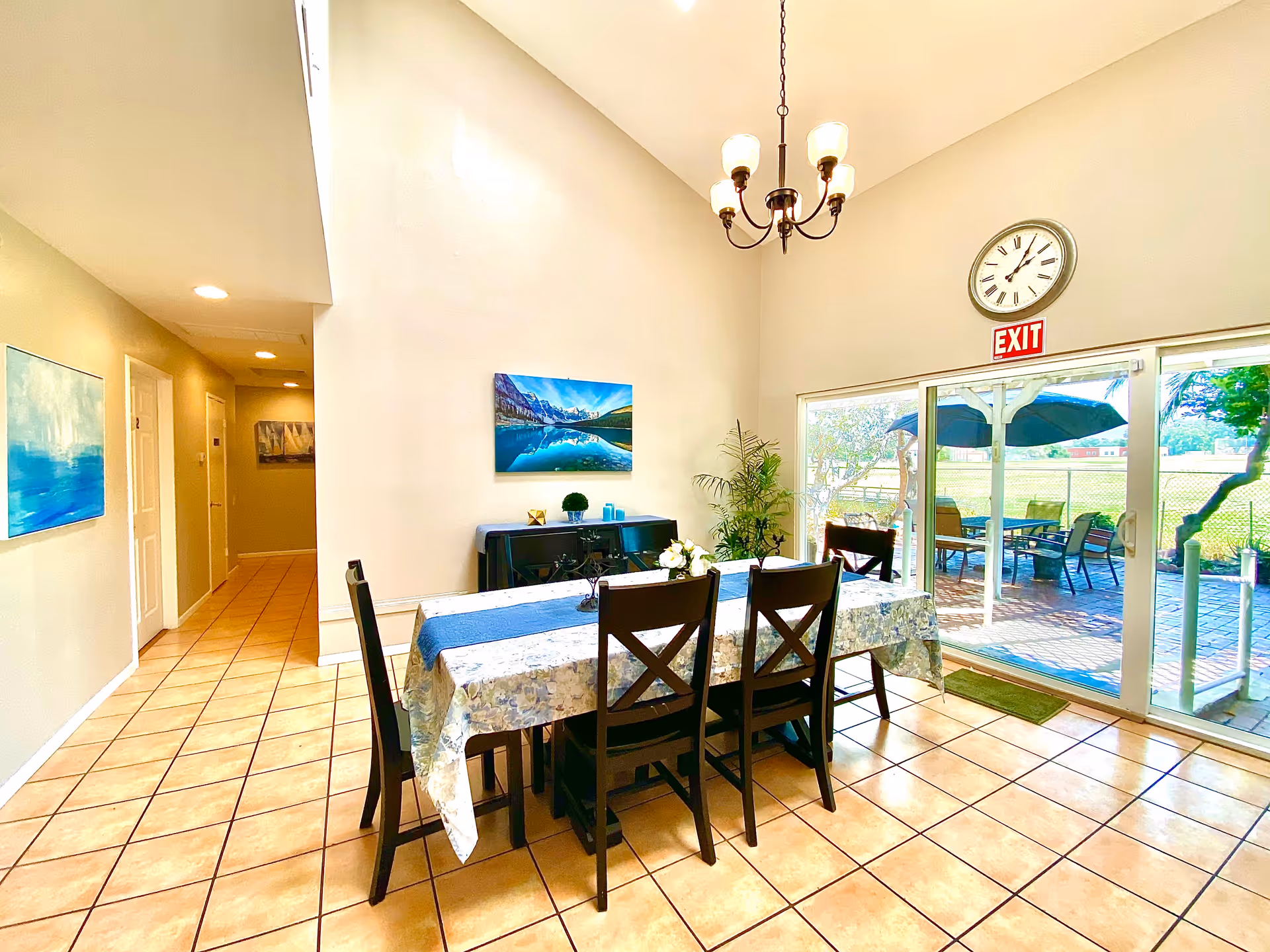 Bright dining area with a rectangular table covered with a floral tablecloth and a blue runner, surrounded by six dark wooden chairs. A chandelier with five lights hangs from the high ceiling. On the wall above a black sideboard is a landscape painting of a lake and mountains. To the right, large sliding glass doors lead to an outdoor patio with tables, chairs, and umbrellas. The floor is tiled, and there are additional paintings on the left wall along a hallway. A clock and an exit sign are mounted above the sliding doors.