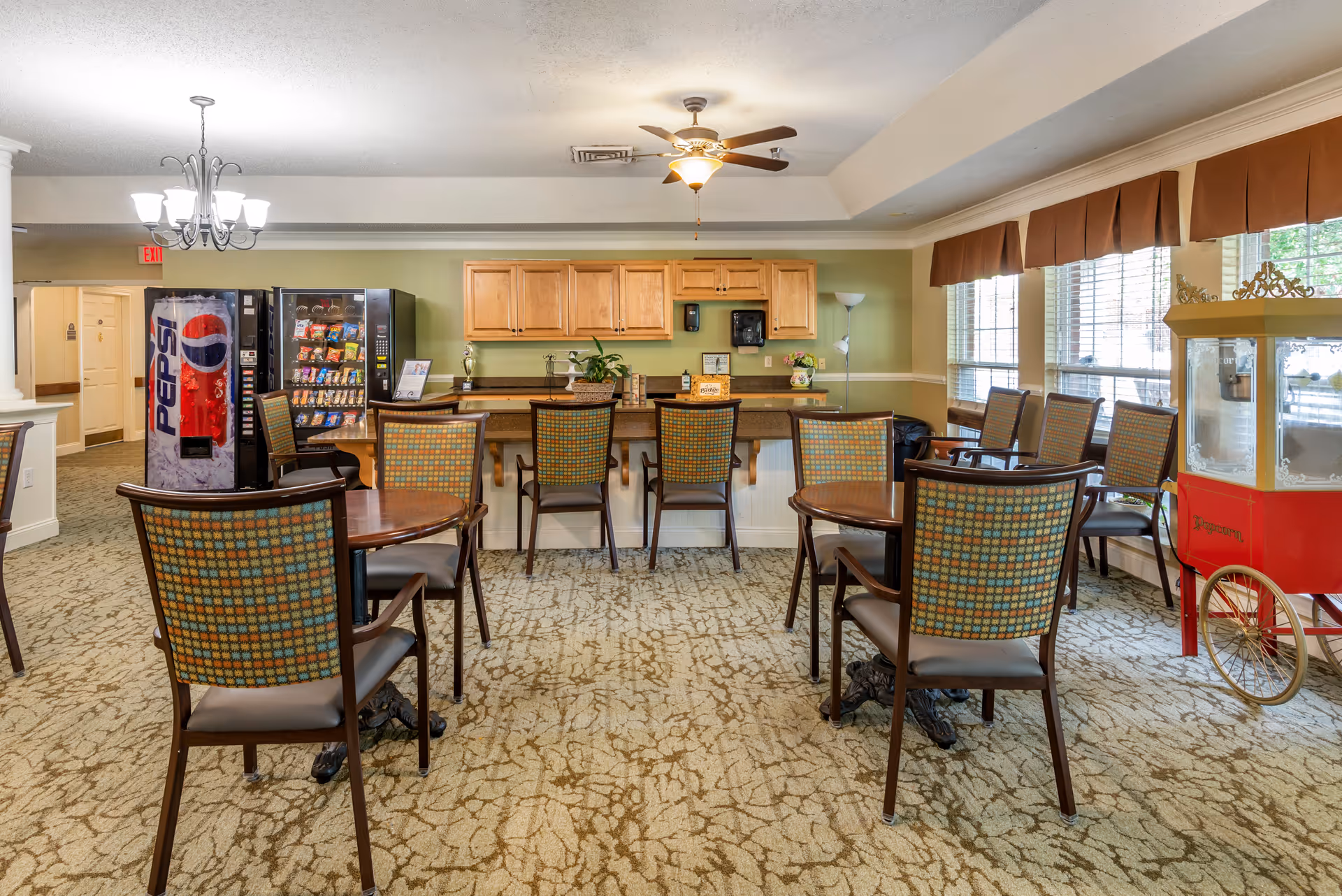 A common area with several round tables and patterned chairs arranged on a carpeted floor. There is a counter with wooden cabinets above it, a Pepsi vending machine, a snack vending machine, and a popcorn machine on wheels near large windows with brown valances. The ceiling has a fan and a chandelier light fixture.