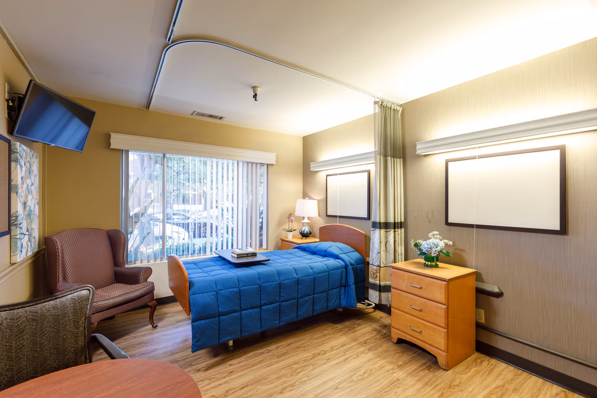 A well-lit senior living bedroom with a single bed covered in a blue quilt, a wooden nightstand with a lamp and a small plant, a wooden dresser with a vase of flowers, two framed wall decorations, a large window with vertical blinds, a maroon armchair, a patterned chair, and a wall-mounted TV.