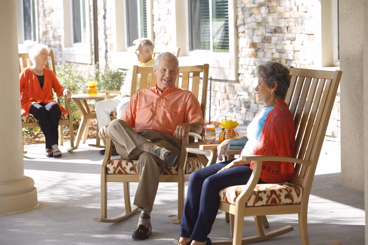 Four elderly people sitting and chatting on a covered patio area with stone walls and large windows. Two are seated in wooden rocking chairs with patterned cushions in the foreground, and two others are seated in the background near a small table with a yellow flower vase.