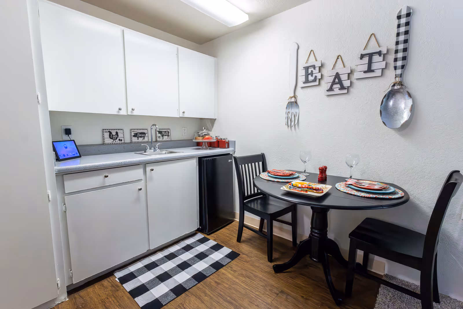 A small kitchen and dining area with white cabinets, a countertop with a sink, a mini refrigerator, and a black round dining table set for two. The wall above the table has decorative hanging letters spelling 'EAT' along with oversized fork and spoon decorations. The floor has a black and white checkered rug in front of the sink.