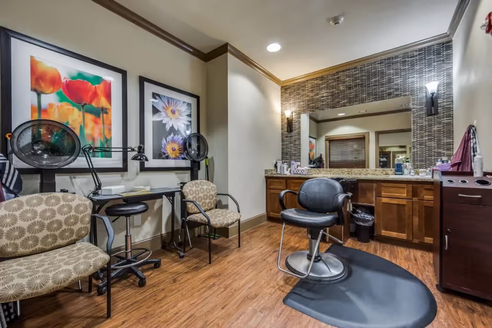 A salon-style grooming room with a styling chair, hooded dryers, seating, a large mirror and floral artwork on the walls.