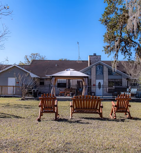 Outdoor view of a single-story assisted living facility building with a patio area covered by a gazebo. In the foreground, there are four wooden Adirondack chairs arranged on a grassy lawn facing the building. The sky is clear and blue.