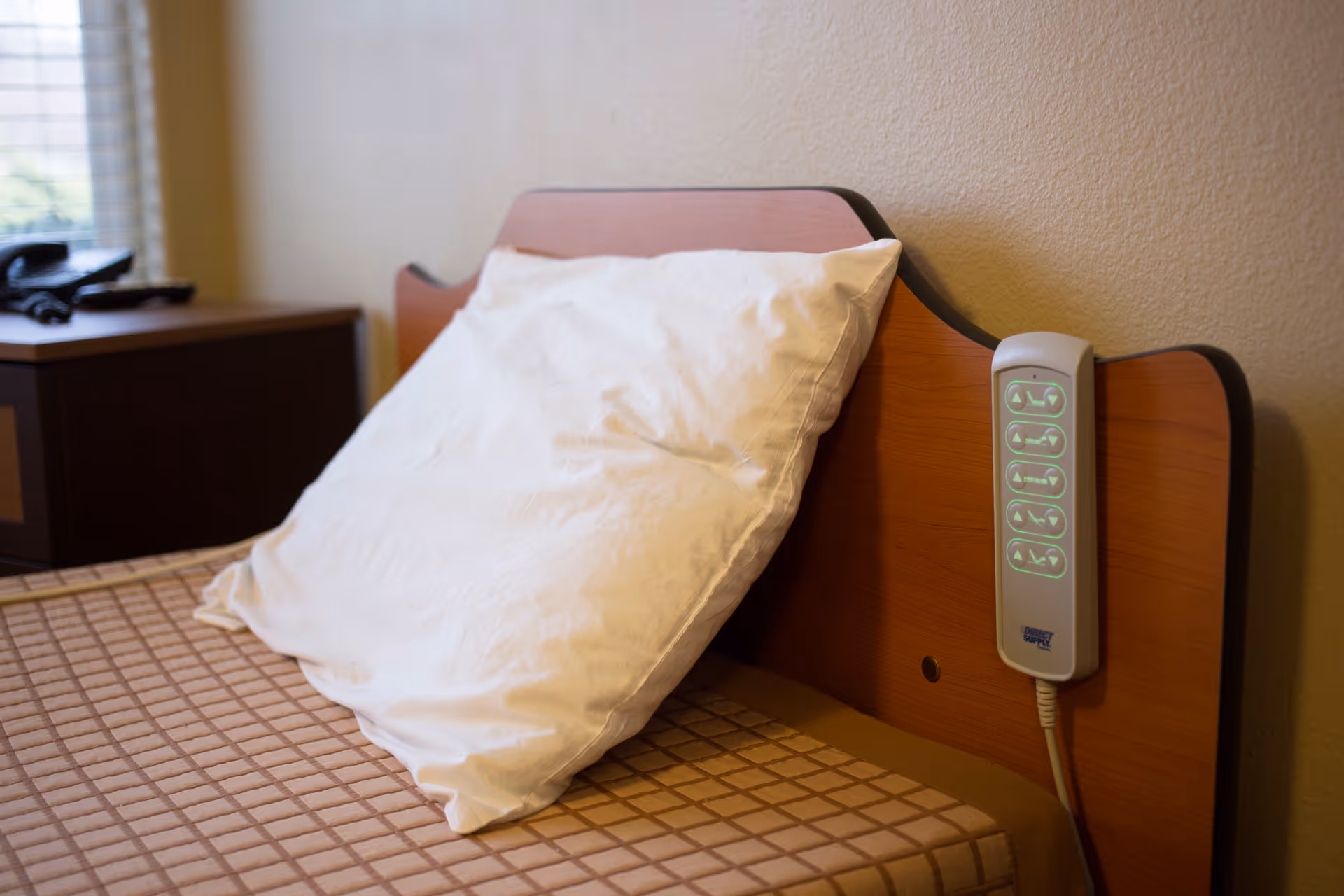 A close-up view of a bed with a white pillow propped up against a wooden headboard. Attached to the headboard is a control panel with illuminated buttons, likely for adjusting the bed position. A nightstand with a telephone is visible in the background near a window with blinds.