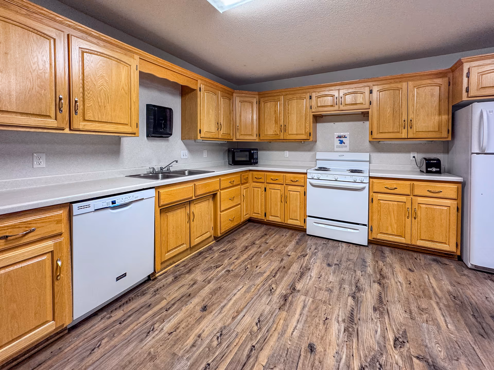 A large kitchen with oak cabinets, white appliances (stove, refrigerator, dishwasher), a double sink, and wood-look flooring.