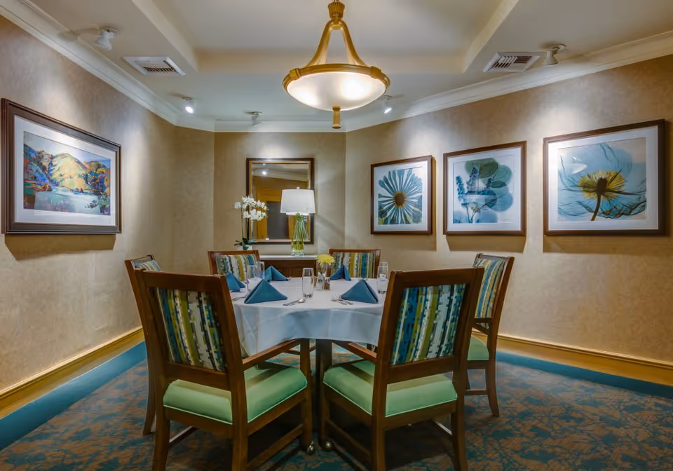A small dining room with a round table set for six people. The table is covered with a white tablecloth and has blue folded napkins, glasses, and silverware. The chairs have wooden frames with green cushioned seats and patterned backs. The walls are decorated with framed artwork featuring floral and landscape themes. A lamp with a white shade and a vase with white flowers sit on a side table against the wall, beneath a mirror. A ceiling light fixture hangs above the table.