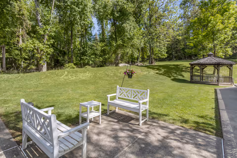 Outdoor patio and lawn with white wooden benches, a small table, a hanging flower basket, and a gazebo near trees.