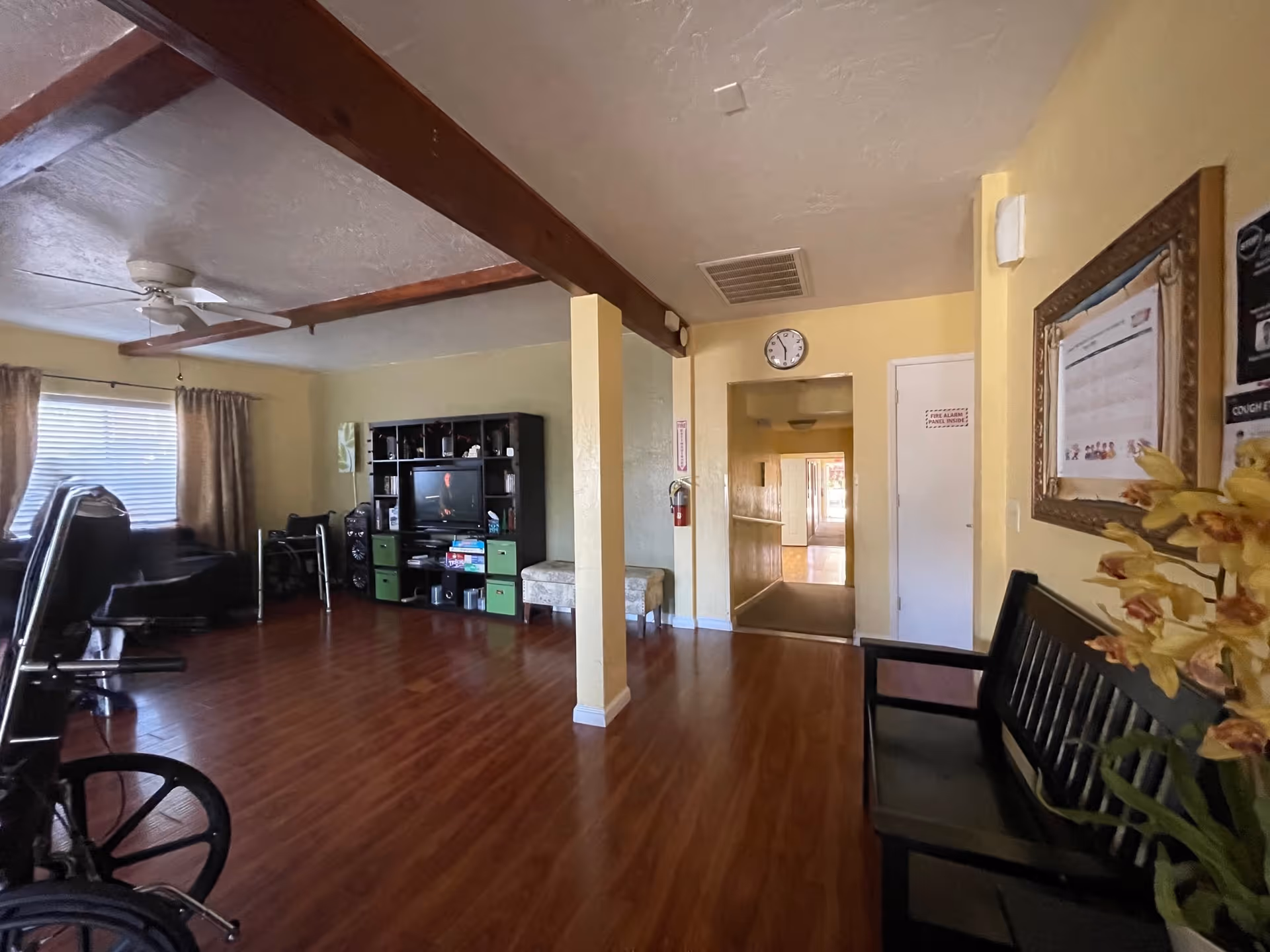 Interior view of a senior living facility common area with wooden flooring, a black entertainment center with a TV, a wheelchair, a walker, a bench with a framed notice above it, and a hallway leading to other rooms. The walls are painted yellow and there is a ceiling fan with wooden beams on the ceiling.