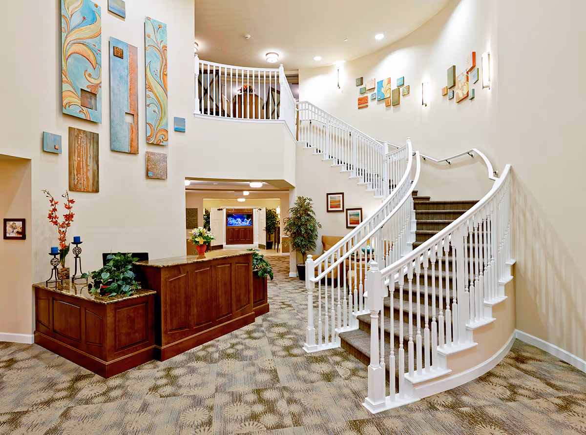 Interior view of a senior living facility featuring a curved staircase with white railings, a wooden reception desk with plants and decorative items, patterned carpet flooring, and colorful abstract wall art. The space is well-lit with wall sconces and ceiling lights, and there is a seating area with a couch and framed pictures on the wall in the background.