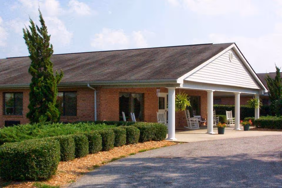 Exterior view of a single-story brick building with a covered porch supported by white columns. The porch has several rocking chairs and hanging plants. The building is surrounded by neatly trimmed bushes and a paved driveway.