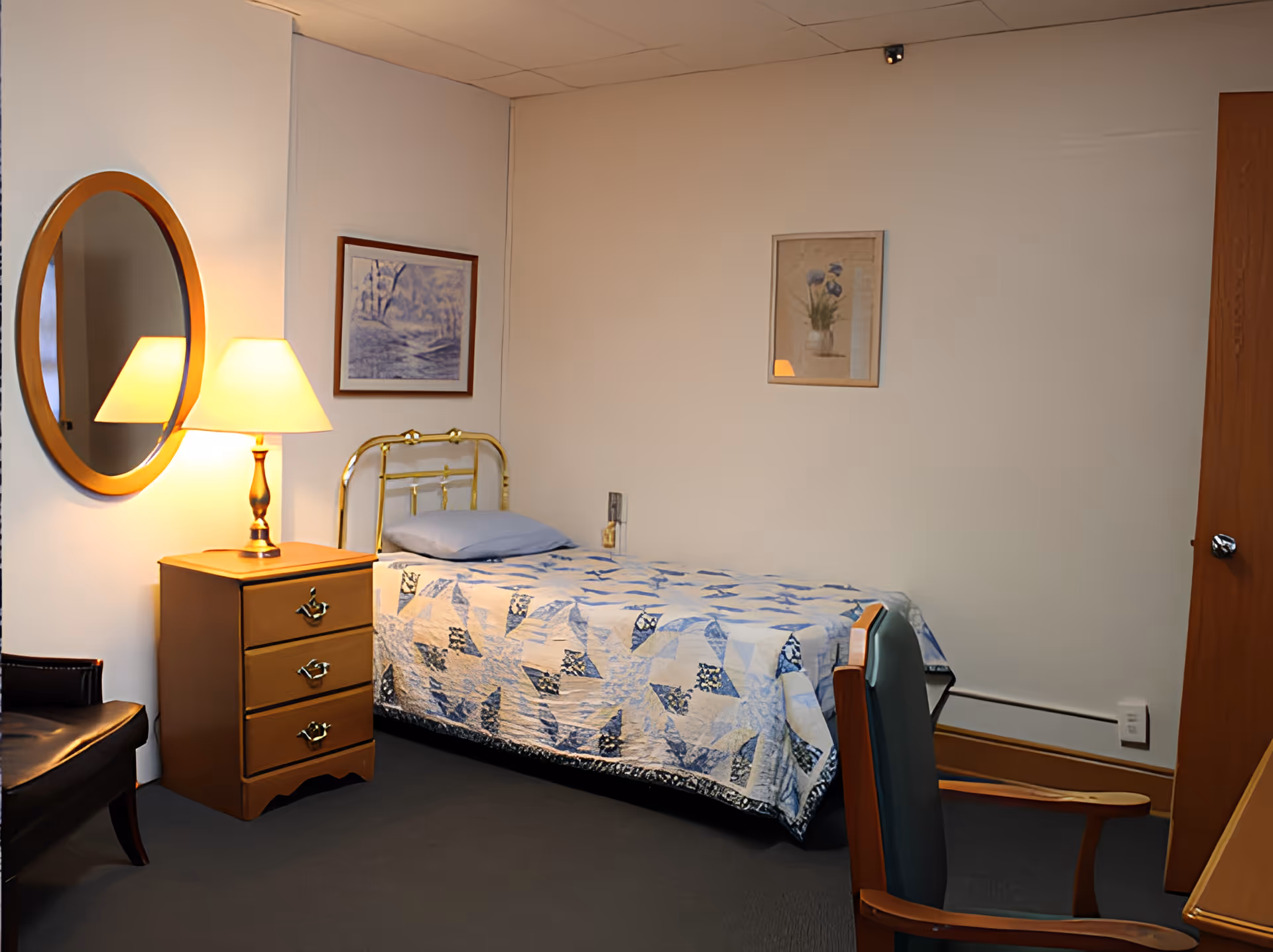Small bedroom with a single bed covered by a blue quilt, a bedside table with lamp, a round wall mirror and chairs.