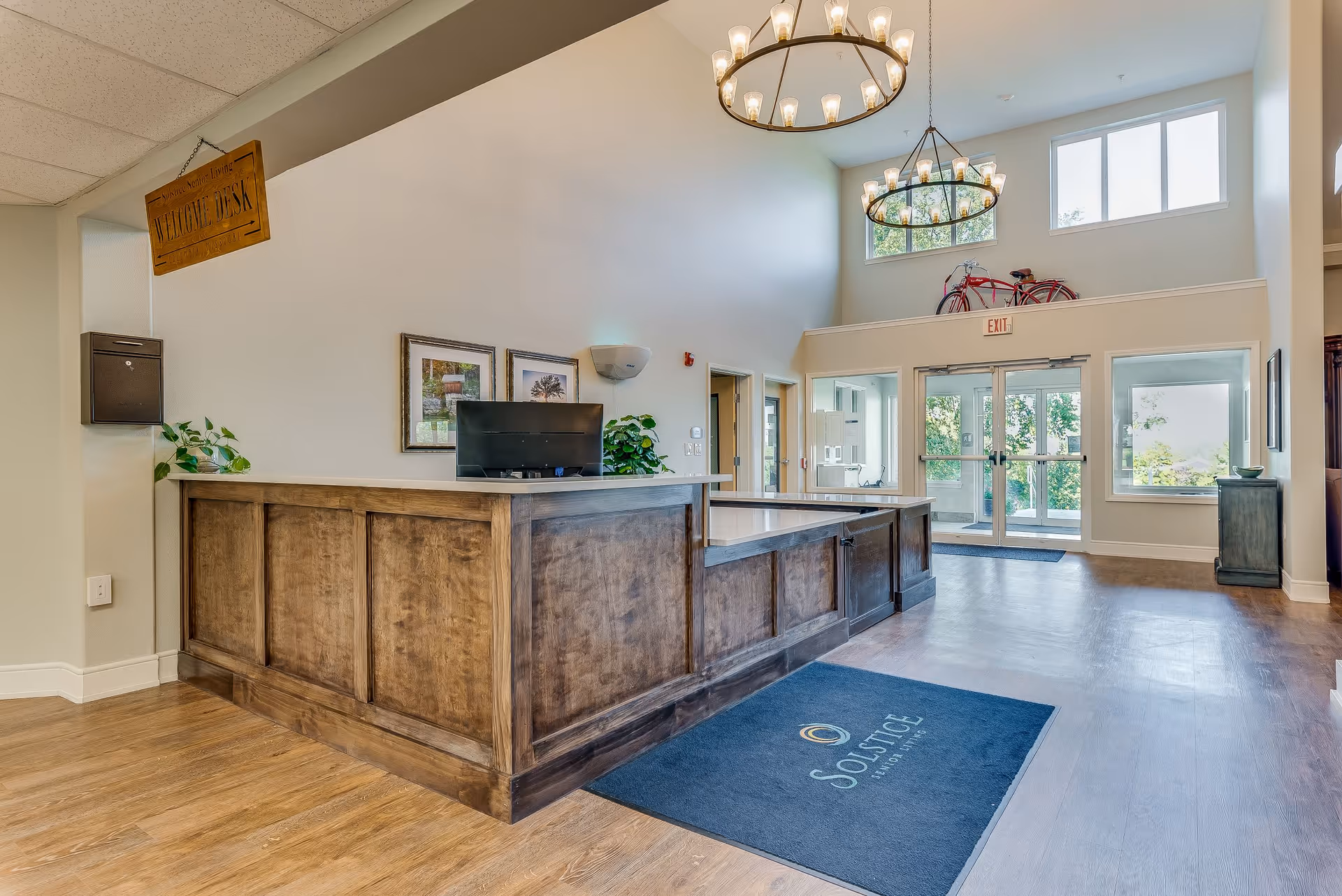 Reception area of Solstice Senior Living at Columbia featuring a wooden front desk with a computer monitor, two framed pictures on the wall, potted plants, large windows letting in natural light, double glass doors leading outside, a blue floor mat with the Solstice logo, and two circular chandeliers hanging from the ceiling.