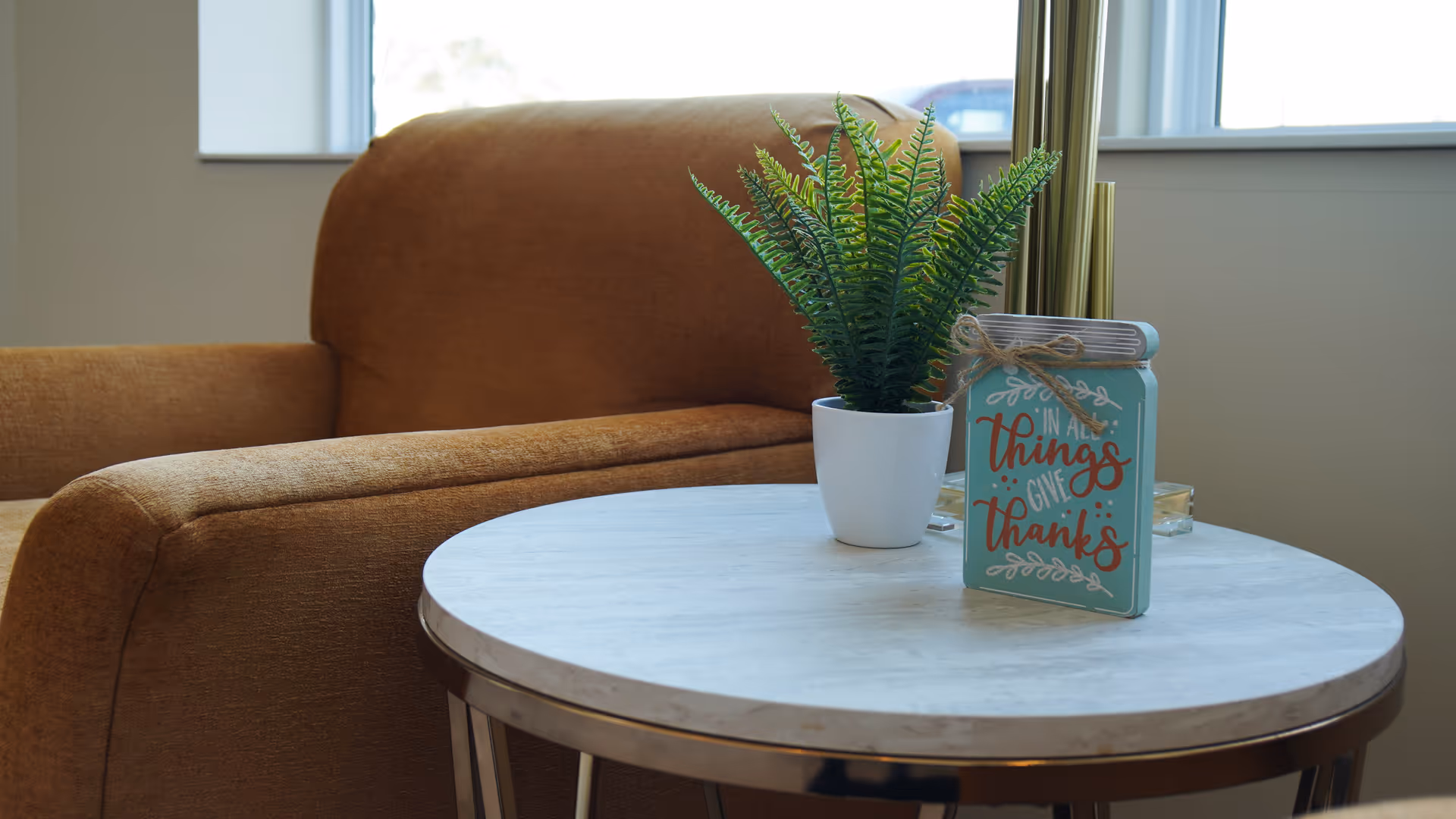 A cozy seating area with a brown upholstered armchair next to a round white marble-top table. On the table, there is a small potted green plant and a decorative sign that reads 'In all things give thanks.' The background shows a window with natural light coming through.