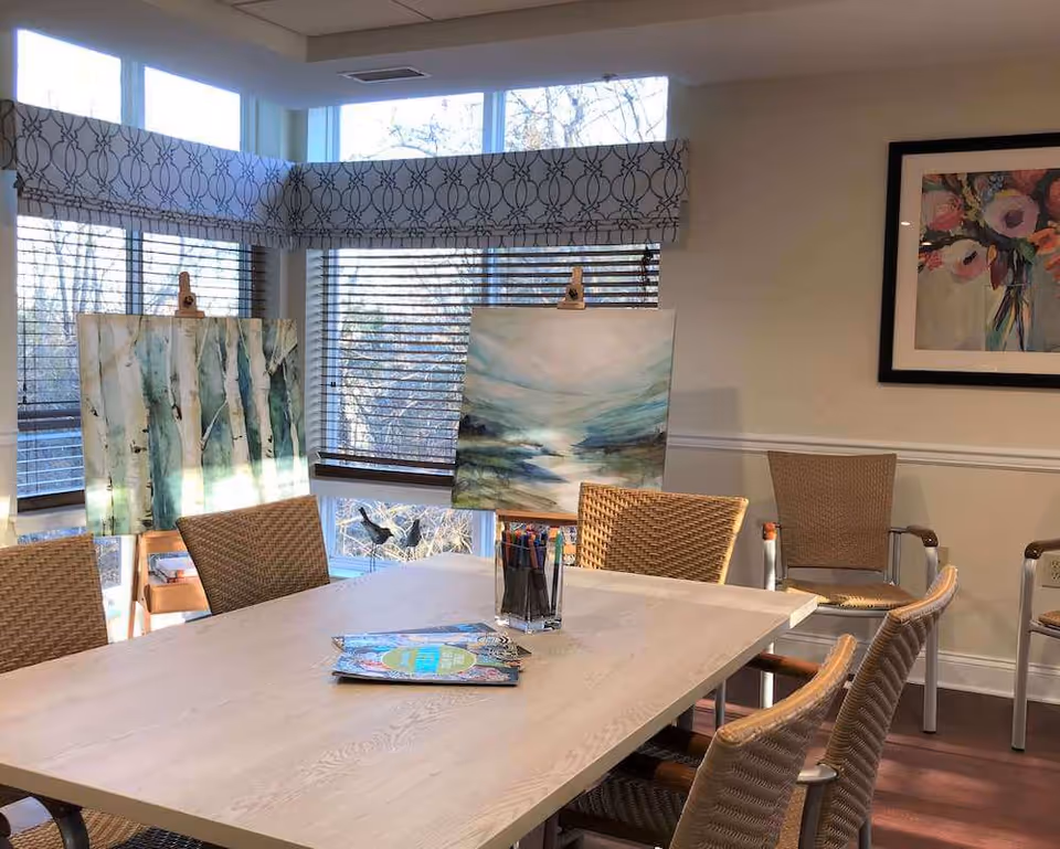 A well-lit room with a rectangular wooden table surrounded by wicker chairs. On the table are coloring books and a container holding colored pencils. Two easels with paintings are positioned near the windows, which have blinds and patterned valances. A framed floral painting hangs on the wall.