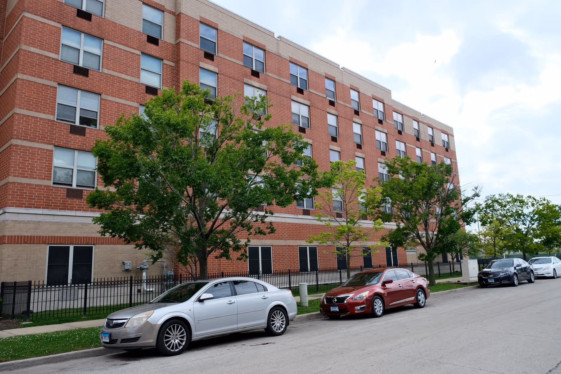 Red-brick, multi-story senior living building front with trees and several cars parked on the street.