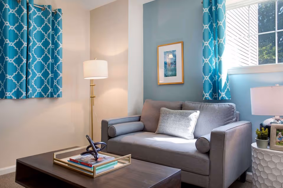 A gray loveseat with pillows in a bright living room featuring teal patterned curtains, a floor lamp, coffee table and window.