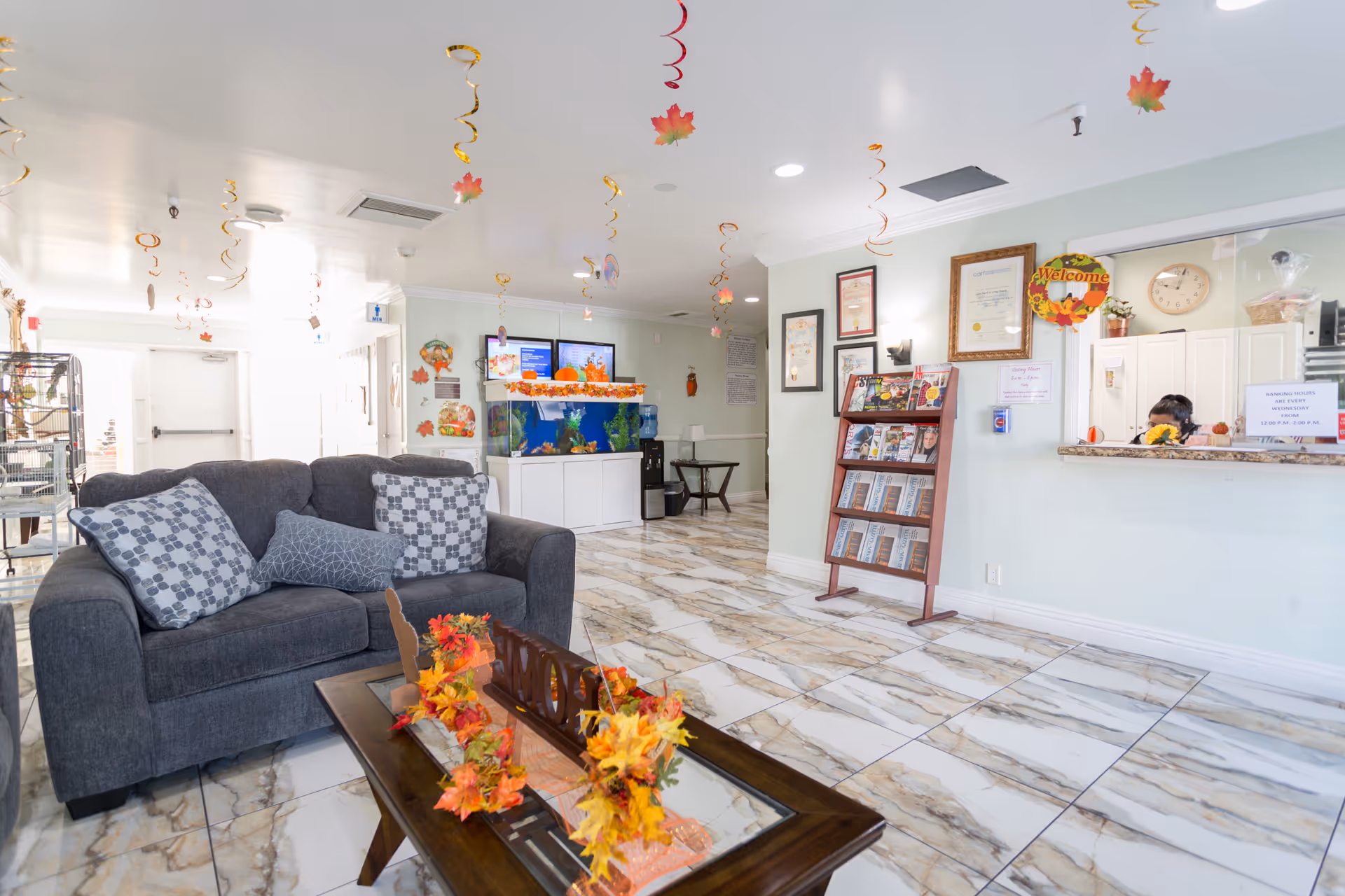 A bright and clean senior living facility lobby with a dark gray couch adorned with patterned pillows, a wooden coffee table decorated with autumn-themed garlands, and a reception desk with a staff member behind it. The floor is tiled with a marble pattern, and the ceiling has hanging spiral decorations with fall leaves. There is a magazine rack and framed certificates on the wall, along with a fish tank and digital screens in the background.
