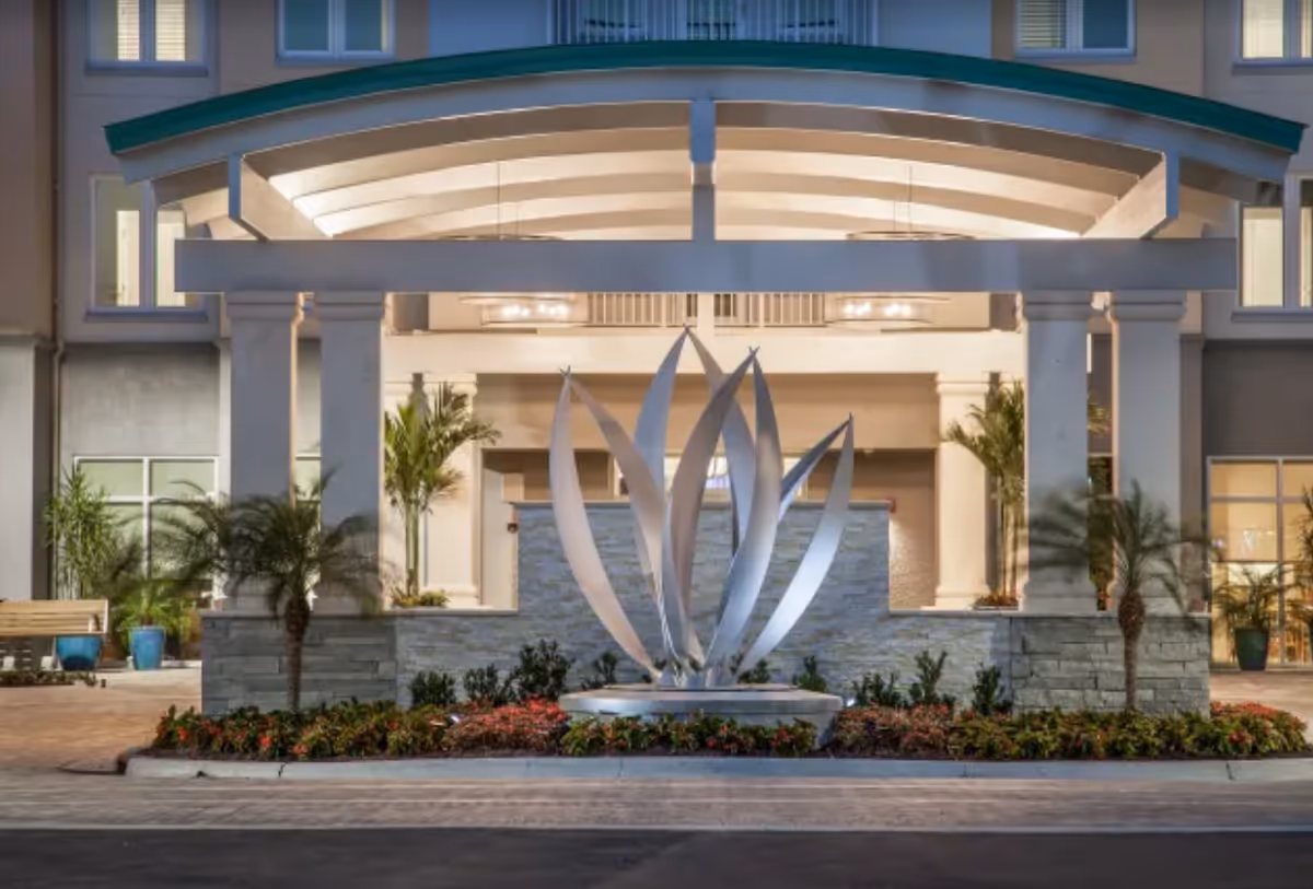 Front entrance of a senior living facility with a modern metal sculpture resembling leaves or petals in the center, surrounded by landscaped plants and flowers. The building features large columns, a covered entryway with ceiling lights, and windows with warm interior lighting.