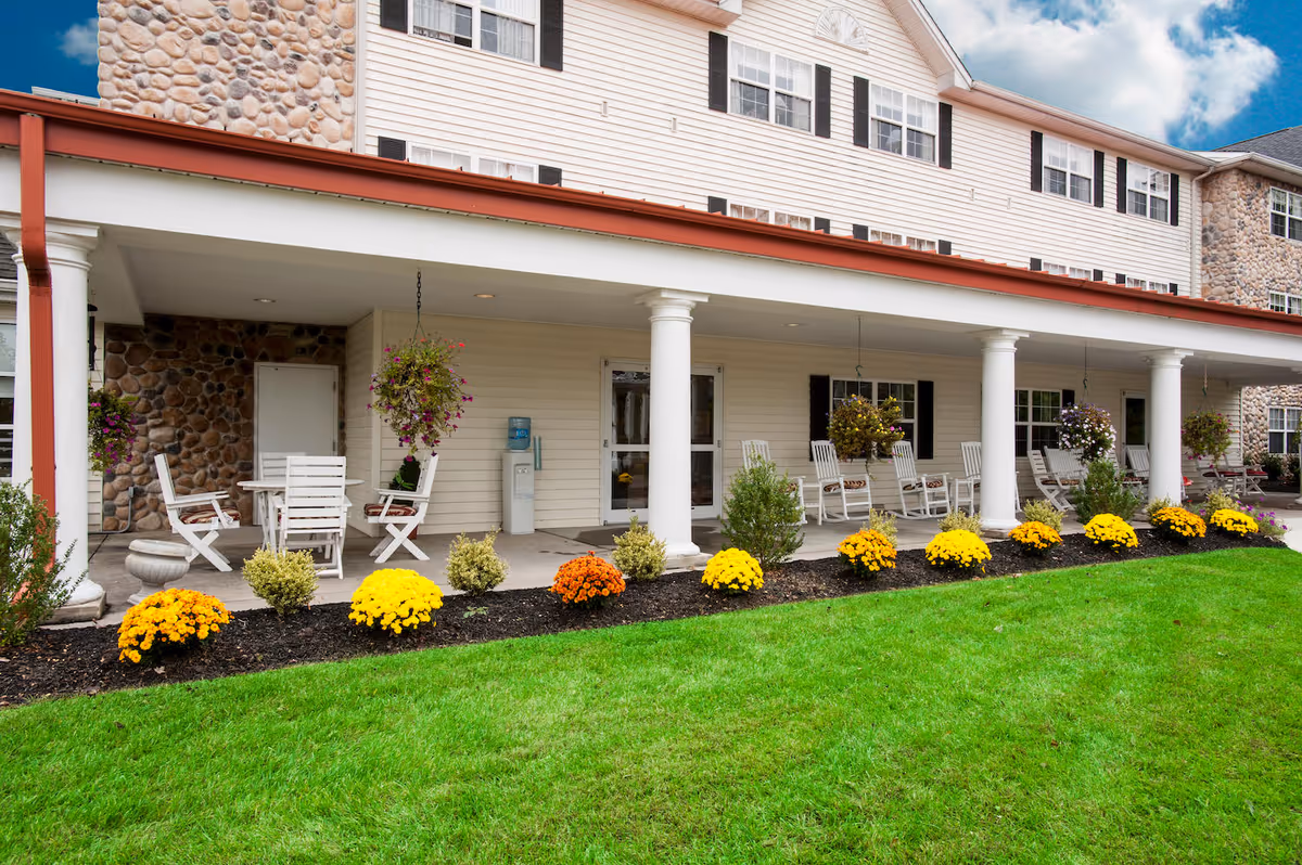 Covered porch area of a senior living facility with white rocking chairs and tables, hanging flower baskets, and a row of yellow and orange flowers planted along the edge of a green lawn. The building exterior features stone and beige siding with white columns supporting the porch roof.