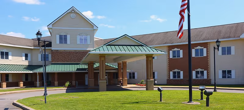 Exterior view of Cooperstown Center, a large multi-story building with beige and brick facade, green metal roofing over the entrance, and an American flag on a flagpole in front. The sky is clear and blue with a few clouds.