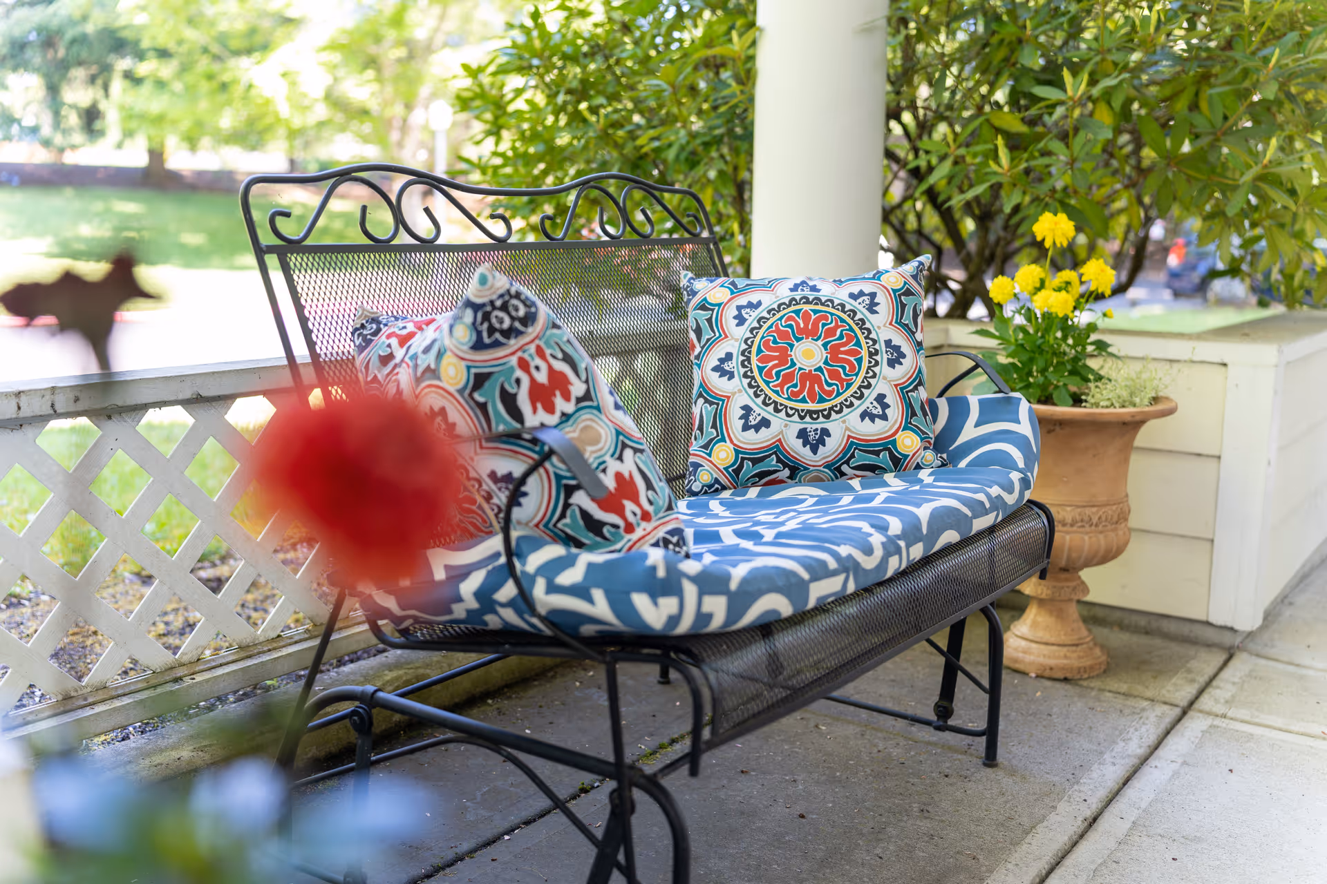 A decorative metal bench with patterned cushions and pillows on a porch next to a potted plant with yellow flowers.