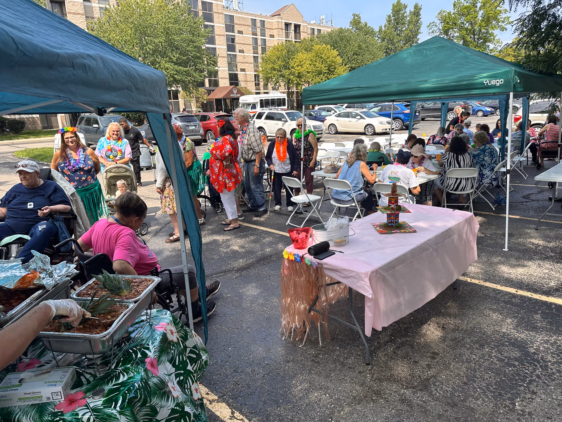 Seniors and staff gathered under pop-up canopies for an outdoor party with tables, a buffet, and a residential building in the background.