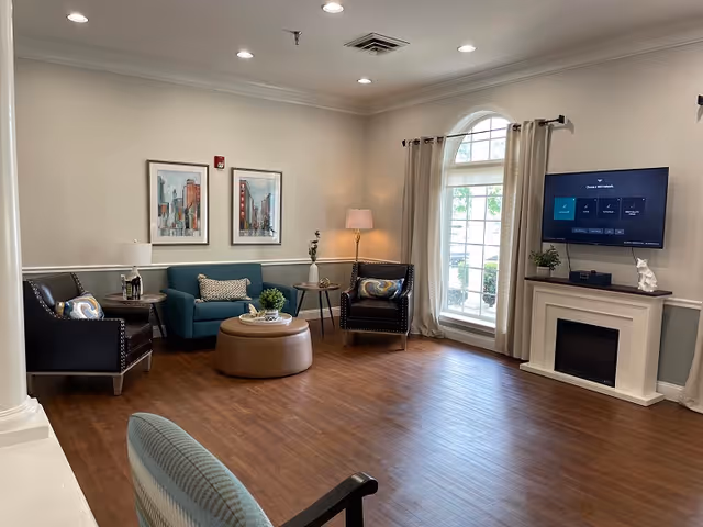 A cozy living room area with two black armchairs and a teal loveseat arranged around a round brown ottoman. There are two framed cityscape paintings on the wall, a floor lamp in the corner, and a large arched window with beige curtains letting in natural light. A flat-screen TV is mounted above a white fireplace mantel with a small plant and decorative item on it. The room has wood flooring and soft, neutral-colored walls.