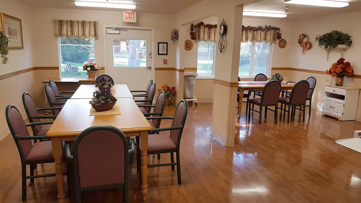 A clean and well-lit dining area in a senior living facility with two wooden tables, each surrounded by six cushioned chairs. The room has polished wooden floors, windows with valances, floral decorations on the walls and tables, and an exit door with a window showing greenery outside.