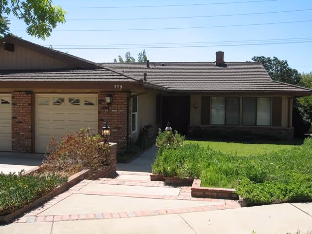 Front exterior view of a single-story residential building with a garage, brick and wood siding, a driveway, and a green lawn with some shrubs and plants.
