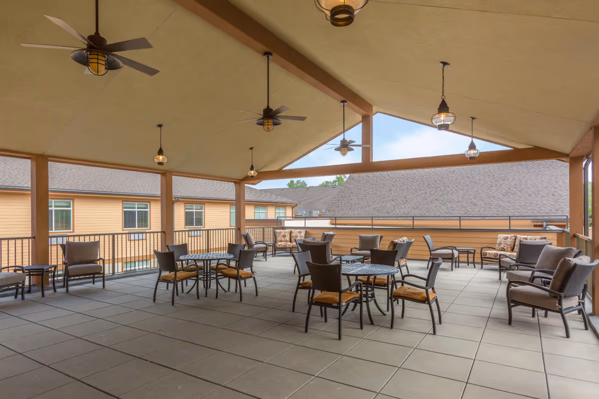 Covered outdoor patio with tables, chairs, ceiling fans and pendant lights overlooking adjacent buildings.