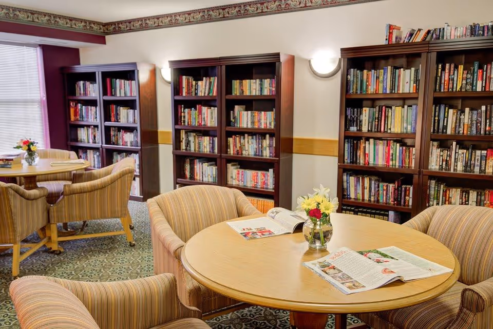 A cozy library room in a senior living facility with multiple wooden bookshelves filled with books. There are round wooden tables surrounded by striped upholstered chairs. On the tables are open magazines and small vases with flowers. The room has patterned carpet and a window with blinds letting in natural light.
