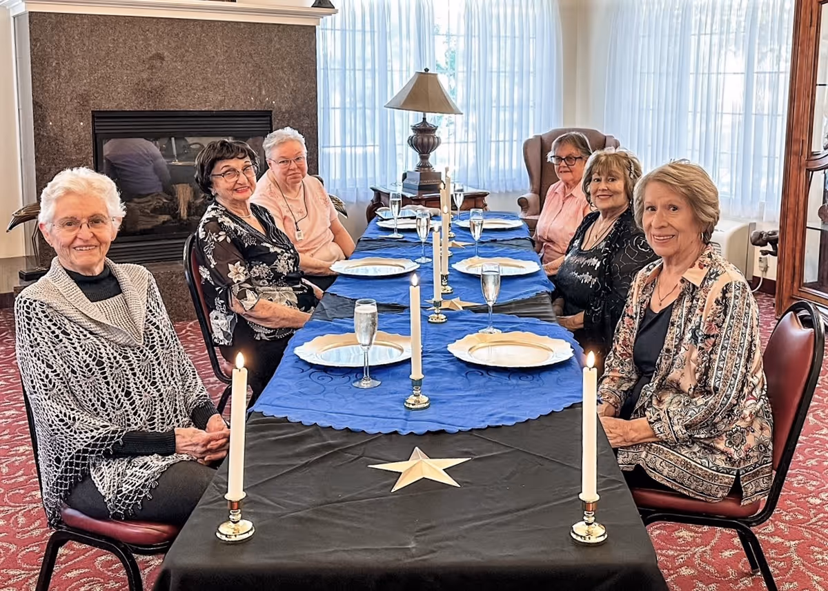 Six elderly women sitting around a long dining table set with plates, glasses, and lit candles in a cozy room with large windows and a fireplace in the background.