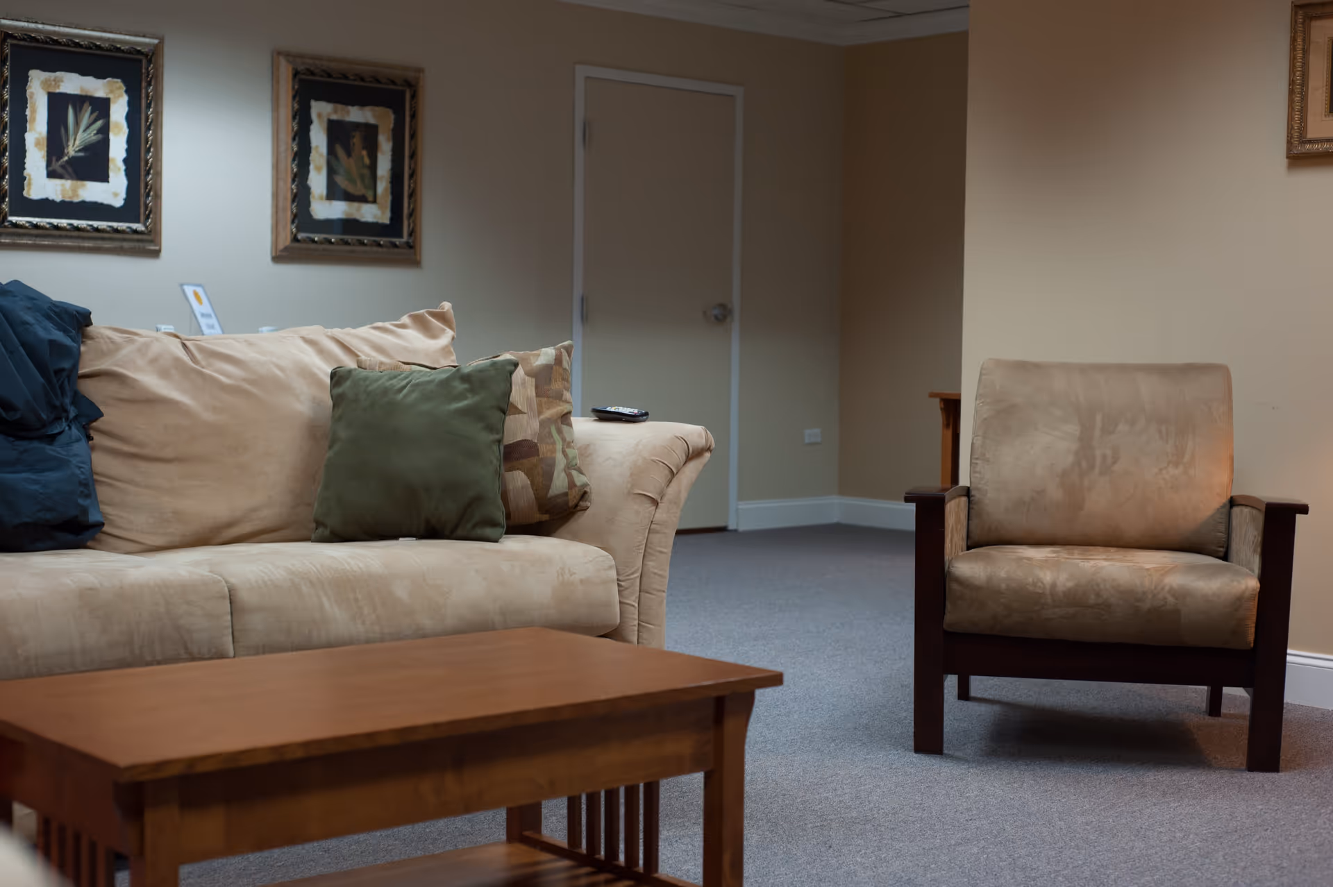 A cozy living room area with a beige couch adorned with green and patterned pillows, a matching beige armchair with wooden armrests, a wooden coffee table in the foreground, and two framed botanical prints hanging on the wall behind the couch.