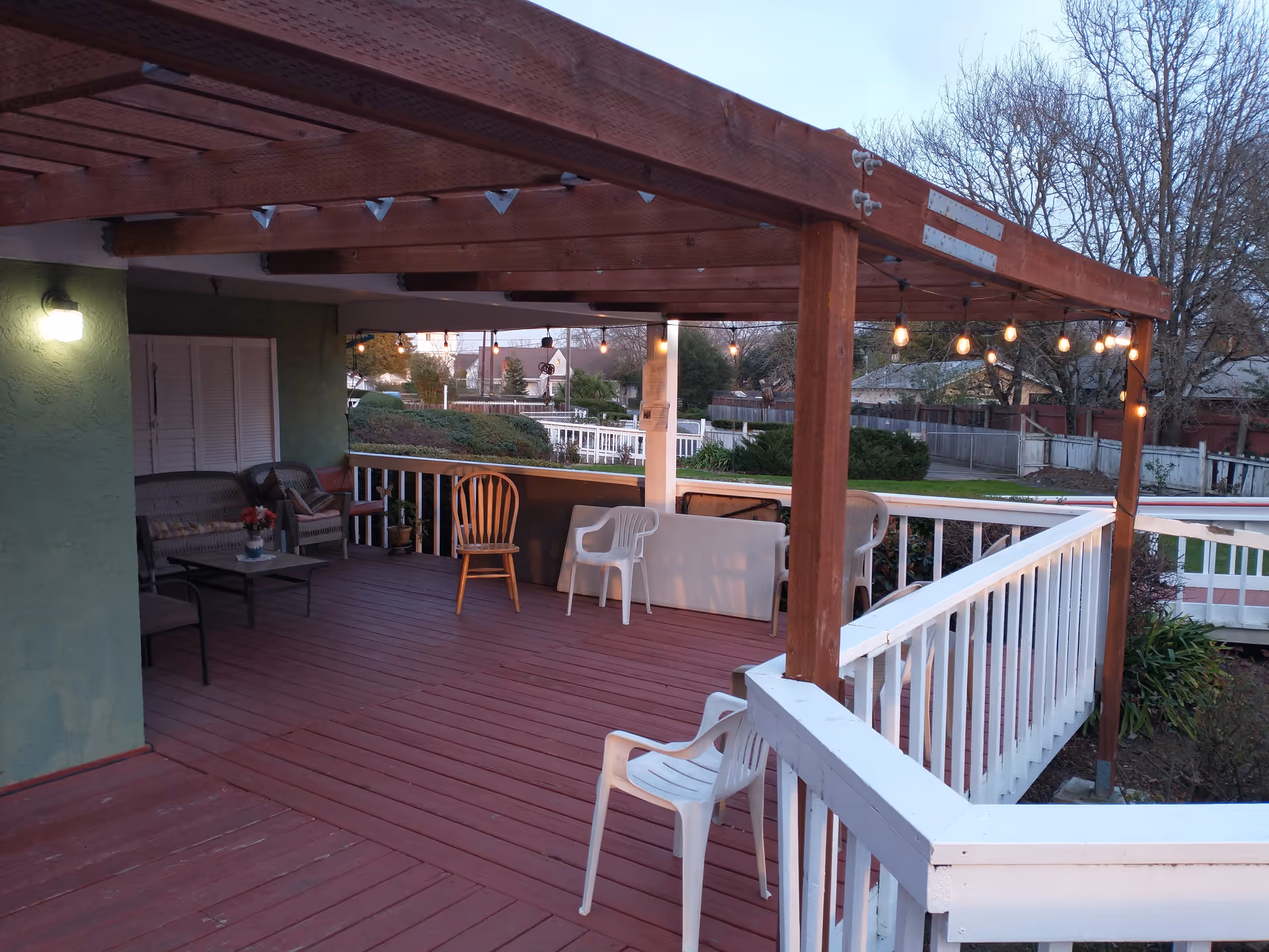A covered outdoor wooden deck with string lights hanging from the wooden beams. The deck has several chairs including plastic and wooden ones, a small table with a flower vase, and a green wall with a light fixture. Surrounding the deck is a white railing, and in the background, there are trees and residential houses.
