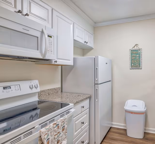 A clean kitchen with white cabinets, a white microwave mounted above a white electric stove, a granite countertop, a white refrigerator, a trash bin, and a decorative wall sign that reads 'Relax you are on BEACH TIME'.