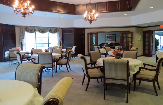 A spacious dining room with multiple round tables covered with white tablecloths, each surrounded by cushioned wooden chairs. The room features large windows with decorative curtains, dark wood paneling, and two ornate chandeliers hanging from the ceiling. Floral centerpieces are placed on the tables, and there are double glass doors leading to another room.